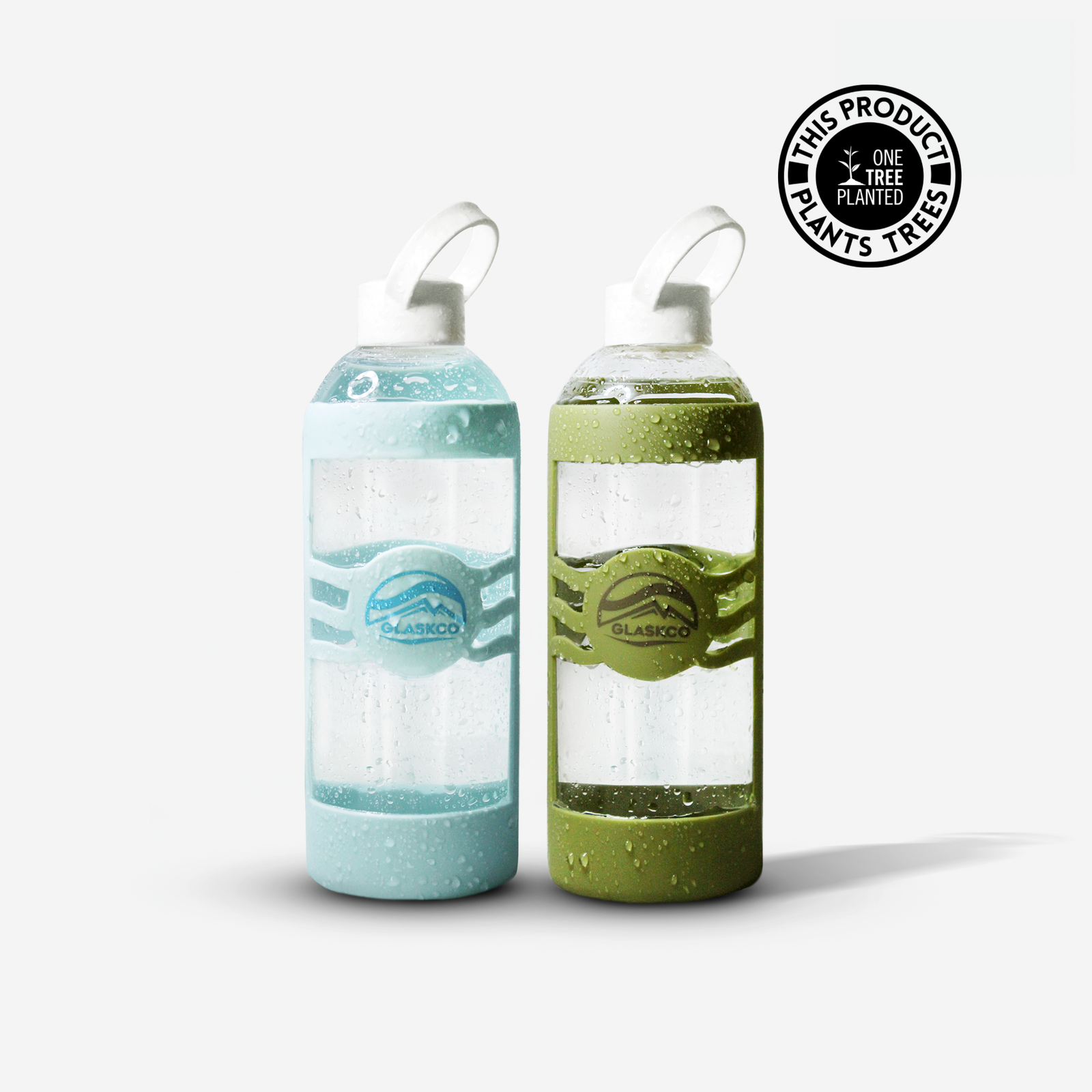 Two elegant Glaskco glass water bottles stand side by side. The bottle on the left boasts a refreshing turquoise gradient while the right one showcases a vibrant green gradient, both adorned with water droplets to emphasize their coolness. Each bottle features a distinct color-matched Glaskco logo and is topped with a white loop handle. A black emblem in the top right states: "This Product Plants One Tree"
