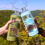 Two hands clinking together Glaskco water bottles in Columbia Blue and Army Green, creating a natural splash effect. Set against a backdrop of sunlit hilly mountains, emphasizing the bottles' outdoor versatility and style.