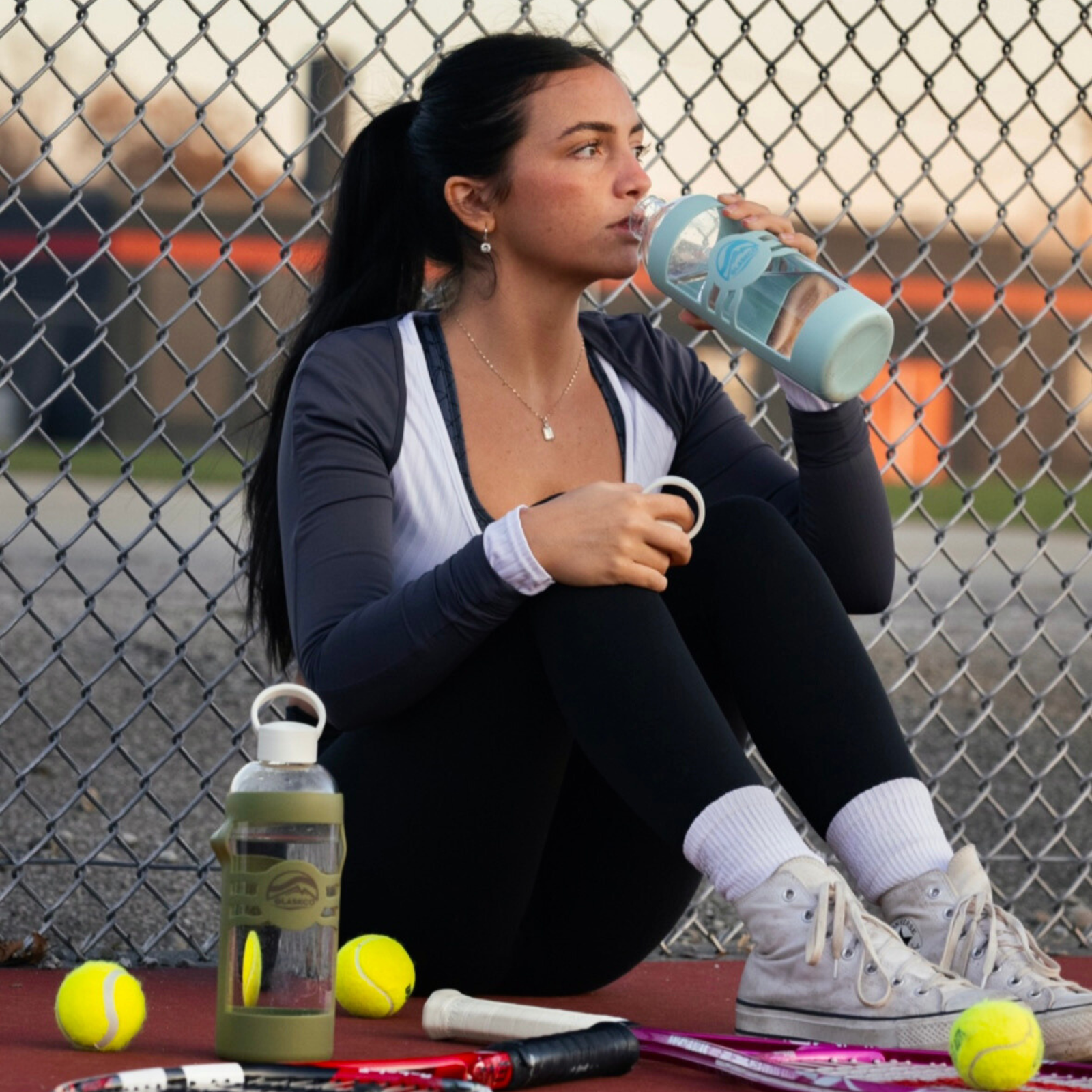 A woman is drinking from a Columbia Blue Glaskco glass water bottle while resting on a tennis court, with an Army Green Glaskco bottle, tennis balls, and a racket in the foreground.