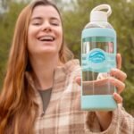 A woman holding a Glaskco glass water bottle with a light blue silicone grip and cap. She is outdoors, wearing a plaid jacket, with greenery in the background.