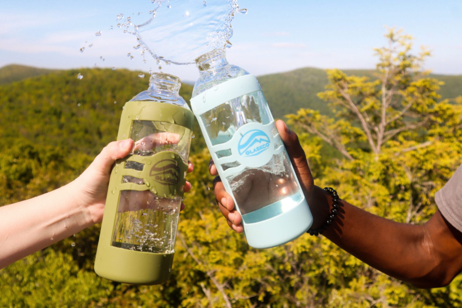 Two hands clinking together Glaskco Water Bottles in Columbia Blue and Army Green, creating a natural splash effect. Set against a backdrop of sunlit hilly mountains, emphasizing the bottles' outdoor versatility and style.