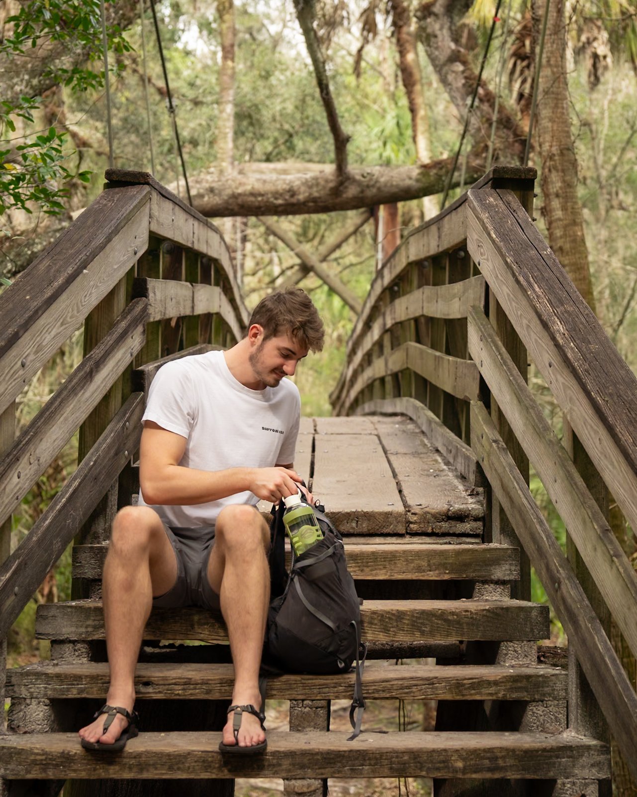 A man sits comfortably on wooden stairs amidst a serene forest setting, captured in the process of retrieving an army green Glaskco water bottle from his backpack.